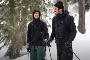 Zwei winterlich gekleidete Personen stehen mit Skistöcken im Schnee und lächeln sich an, umgeben von verschneiten Bäumen.