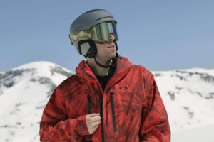 Eine Person, die eine rote Skijacke, einen Helm und eine Skibrille trägt, steht im Freien mit schneebedeckten Bergen im Hintergrund.