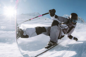 Ein Skifahrer in Winterausrüstung und Helm macht eine scharfe Kurve auf einer verschneiten Piste mit Bergen und hellem Sonnenlicht im Hintergrund.