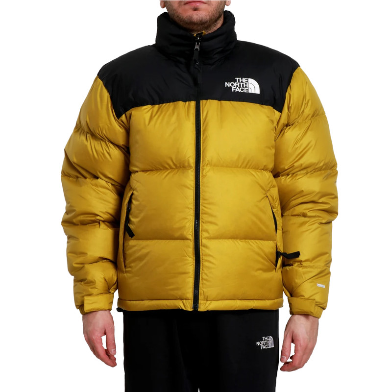 Eine Person, die die Jacke The North Face M 1996 Retro Nuptse Jacket in Deep Dijon/TNF Black mit Reißverschlusstaschen trägt, steht vor einem schlichten weißen Hintergrund.