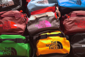 Eine Vielzahl von The North Face Seesäcken in verschiedenen Farben, darunter grün, gelb, braun und rot, sind dicht nebeneinander gestapelt.