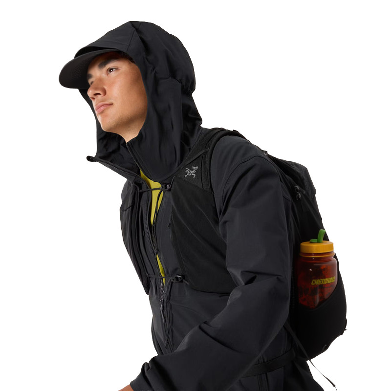 Eine Person in schwarzer Kapuzenjacke und Mütze trägt einen schwarzen Rucksack mit einer Wasserflasche in der Seitentasche und blickt nach links auf einen weißen Hintergrund.